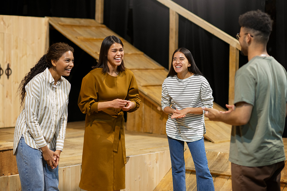 A group of people in street clothes laugh onstage at a theater